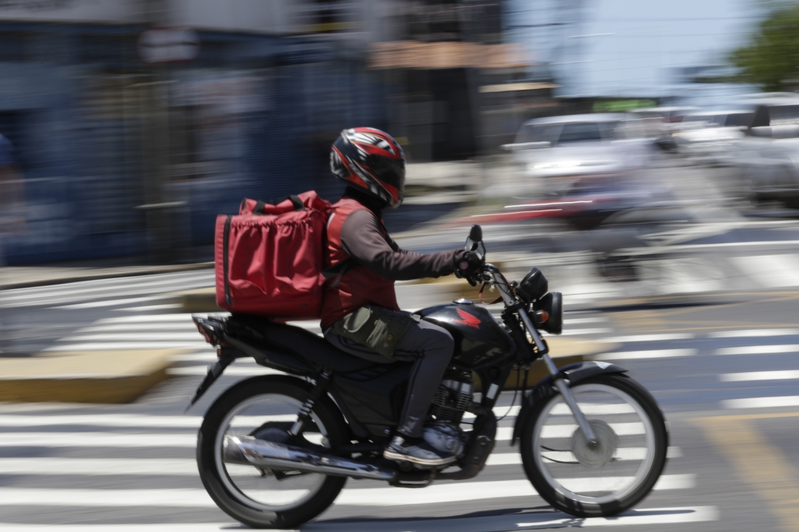 Imagem mostra motoboy na correria em rua movimentada.